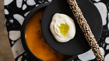 Un bol de soupe garni d’une cuillerée de crème et accompagné d’un bâtonnet de pain saupoudré de graines de sésame, le tout présenté sur une assiette noire posée sur un sol en terrazzo.