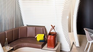 A modern hotel room with a curved brown sofa, a small yellow bag, a brown leather bag, and a white chair. - OKKO Hotels Bayonne Centre (Bayonne)