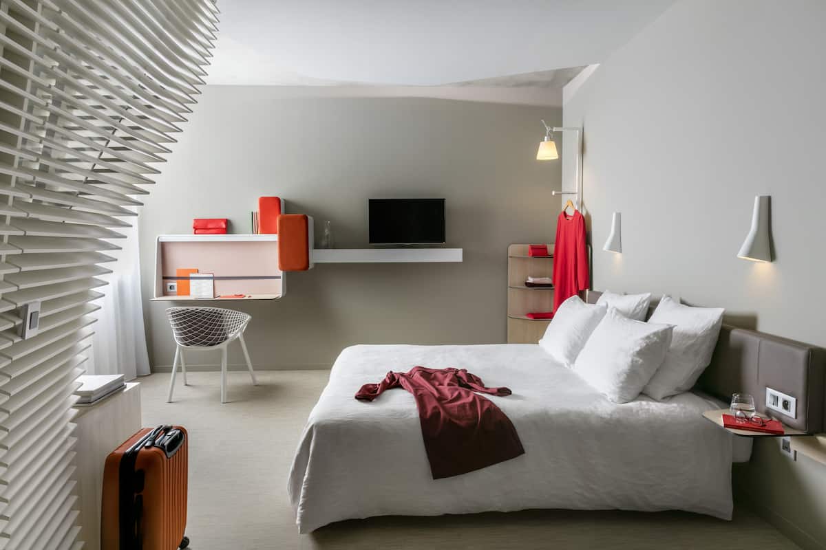 Moderni hotellihuone, jossa on suuri sänky, työpöytä ja televisio sekä oleskelualue, jossa on tuoli.