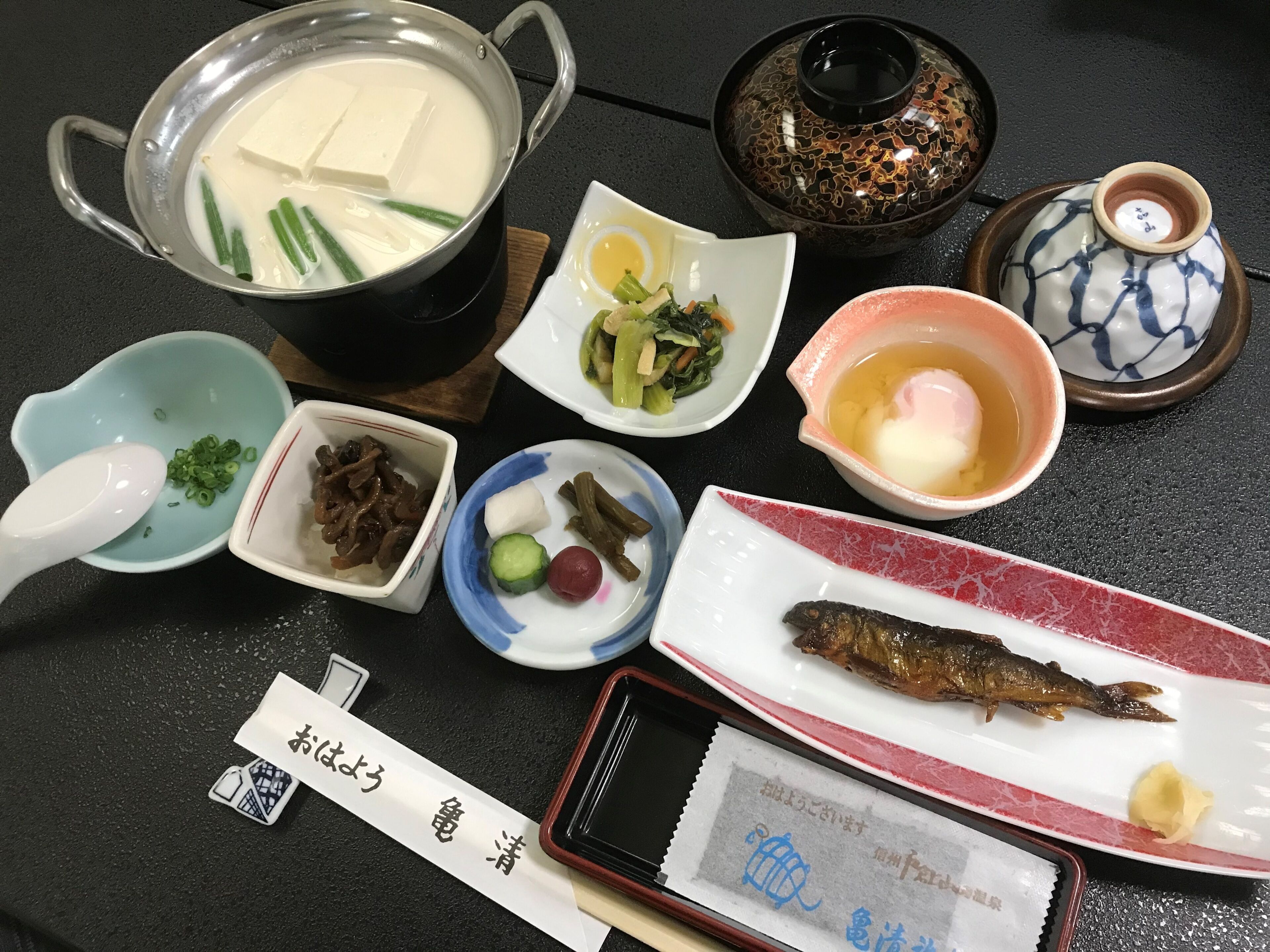 A table with various dishes, including a pot of soup, a bowl of vegetables, a plate of fish, and a dish with tofu.