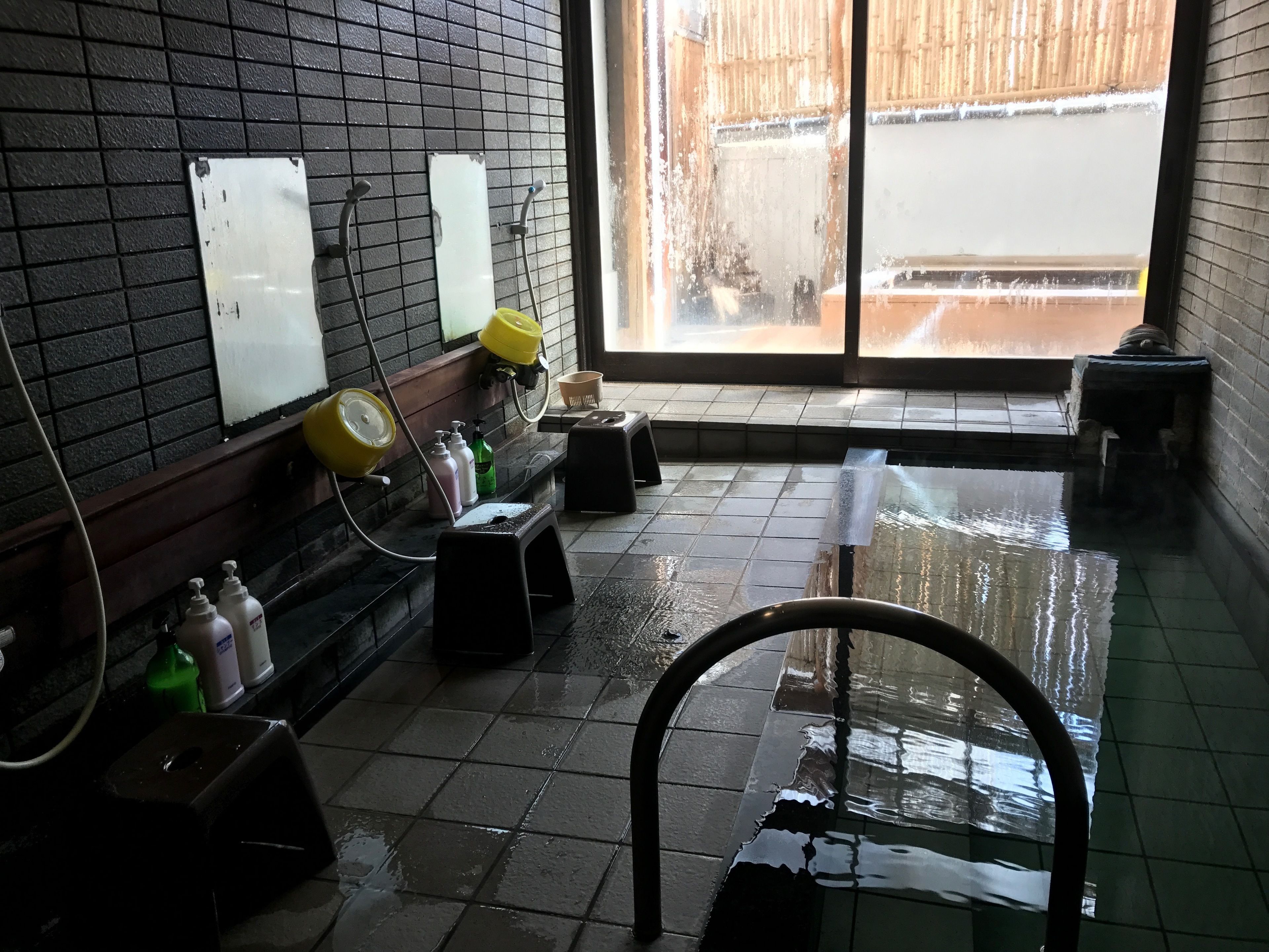 A wet bathroom with a tiled floor, a yellow shower head, and a window with a view of a wooden fence.