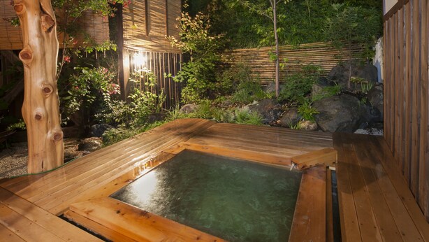 A wooden hot spring bath surrounded by a bamboo fence and lush greenery.