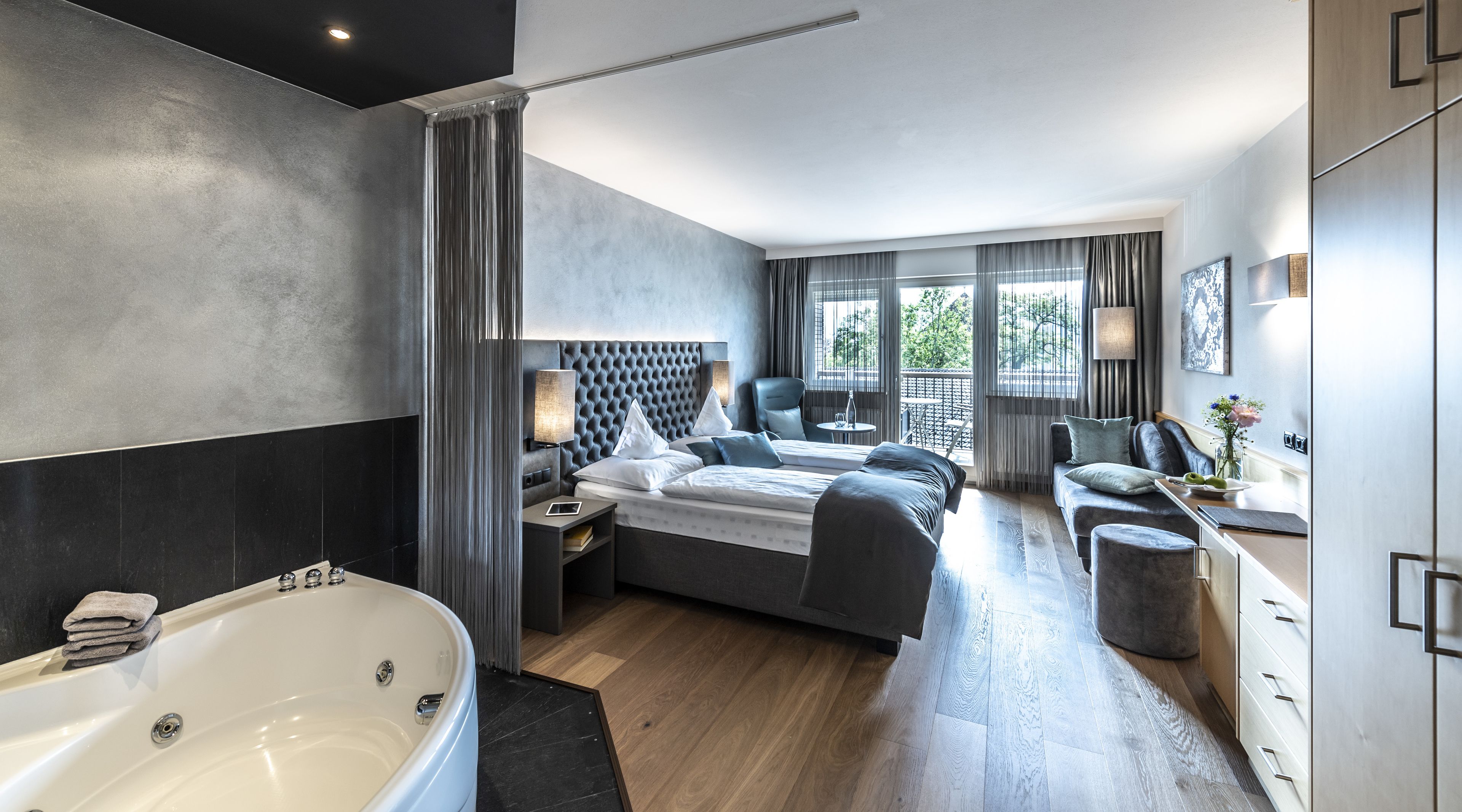 A modern hotel room with a large bed, a separate seating area, a bathtub, and a view of the outdoors.