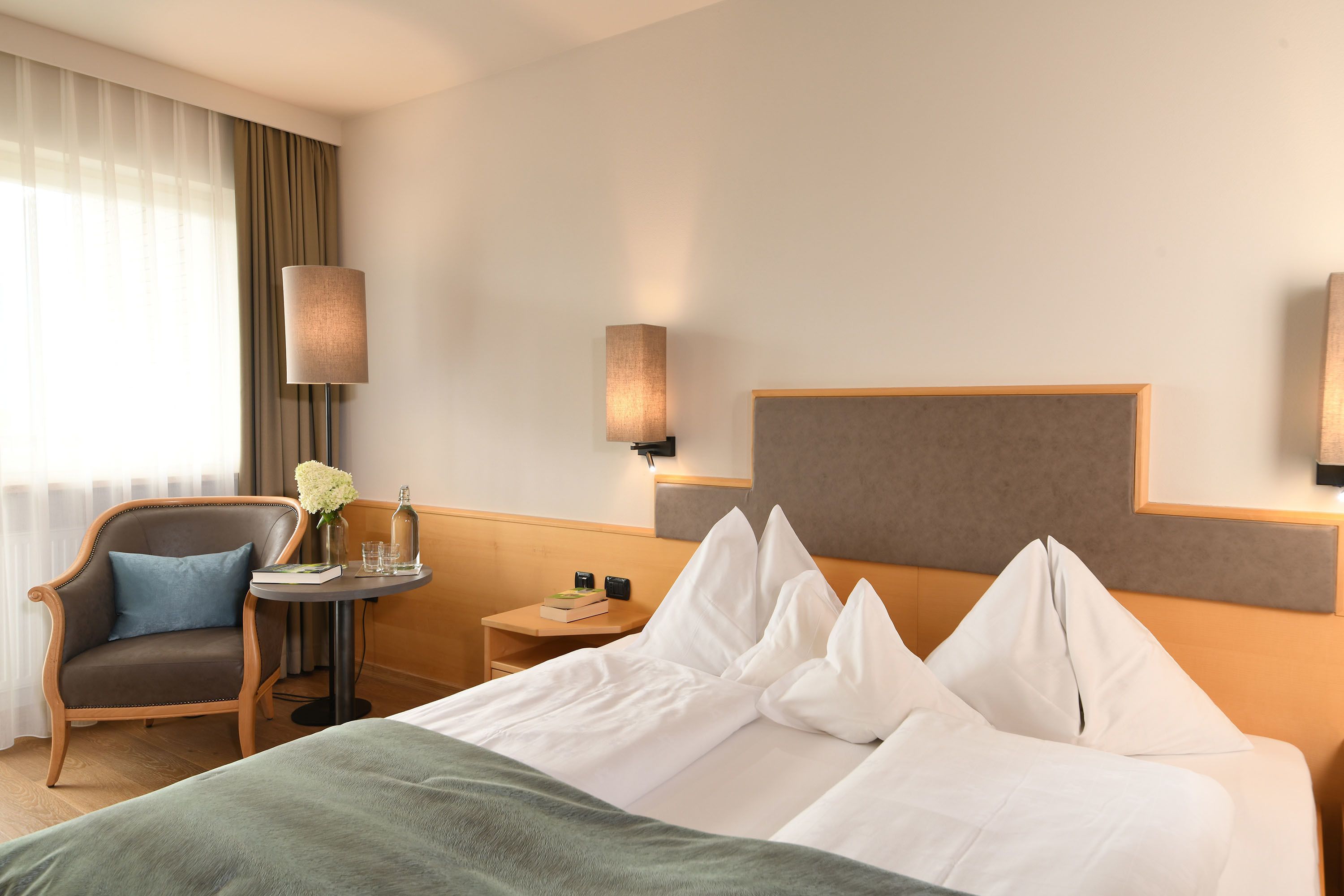 Ein Hotelzimmer mit einem Bett, einem Sessel, einem kleinen Tisch mit Blumenvase und zwei Wandlampen.