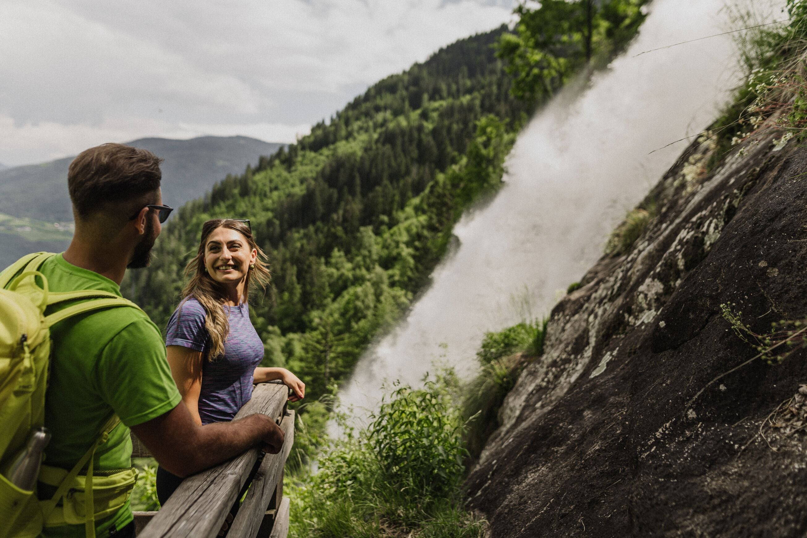 A couple standing on a wooden bridge overlooking a river with a forested hillside in the background.