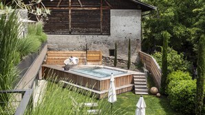 Un chalet rustique doté d’un bain à remous et d’un espace extérieur aménagé, entouré d’une végétation luxuriante et avec une vue sur les montagnes en arrière-plan.