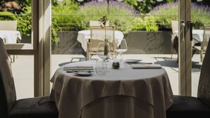 Un espace de restauration en plein air avec une table dressée pour deux personnes, entourée de chaises, et une vue sur la verdure et les montagnes, visible à travers une porte vitrée.