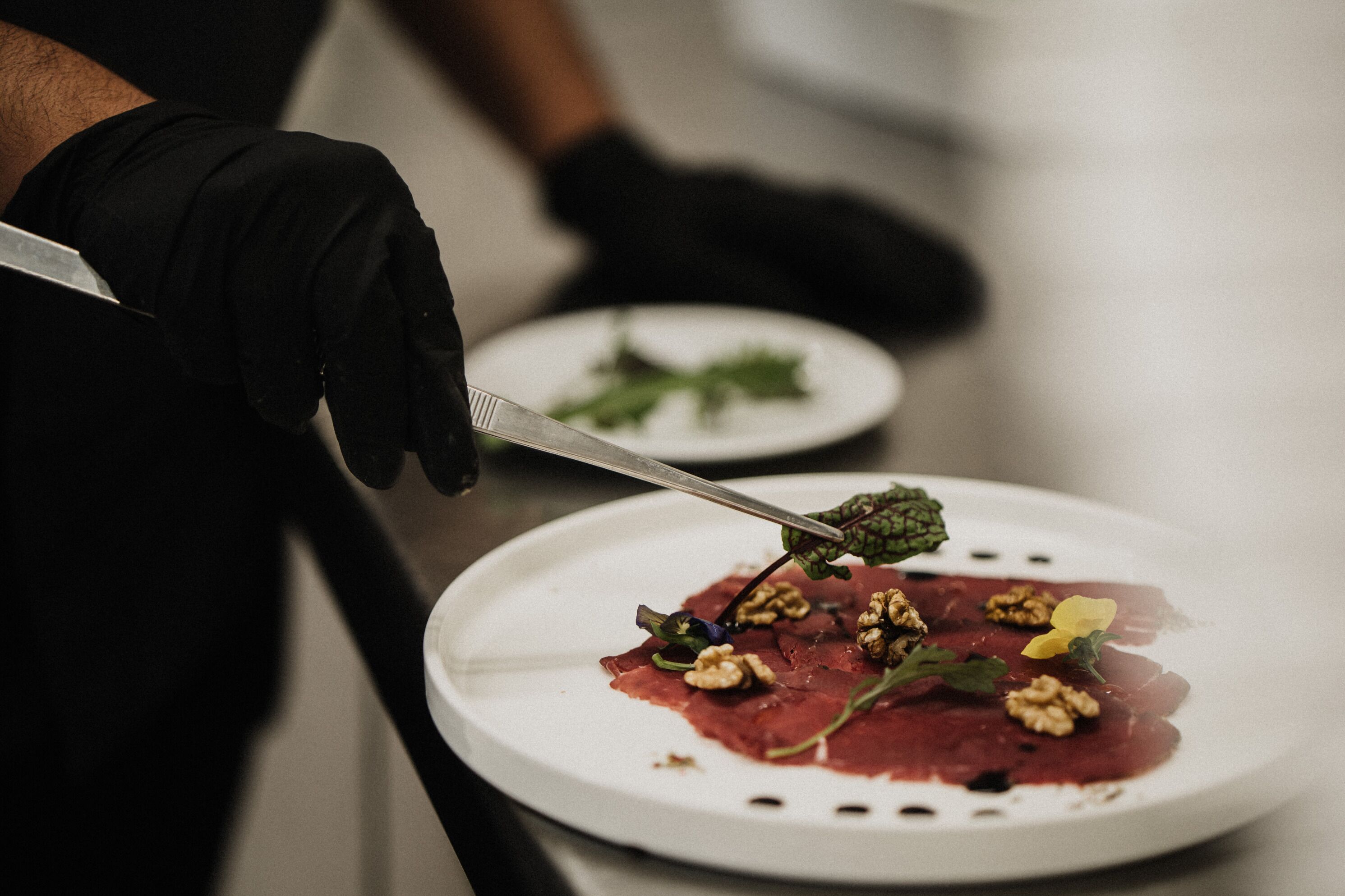 En person med svarta handskar håller i en kniv och förbereder en rätt med kött, valnötter och grönsaker på en vit tallrik.