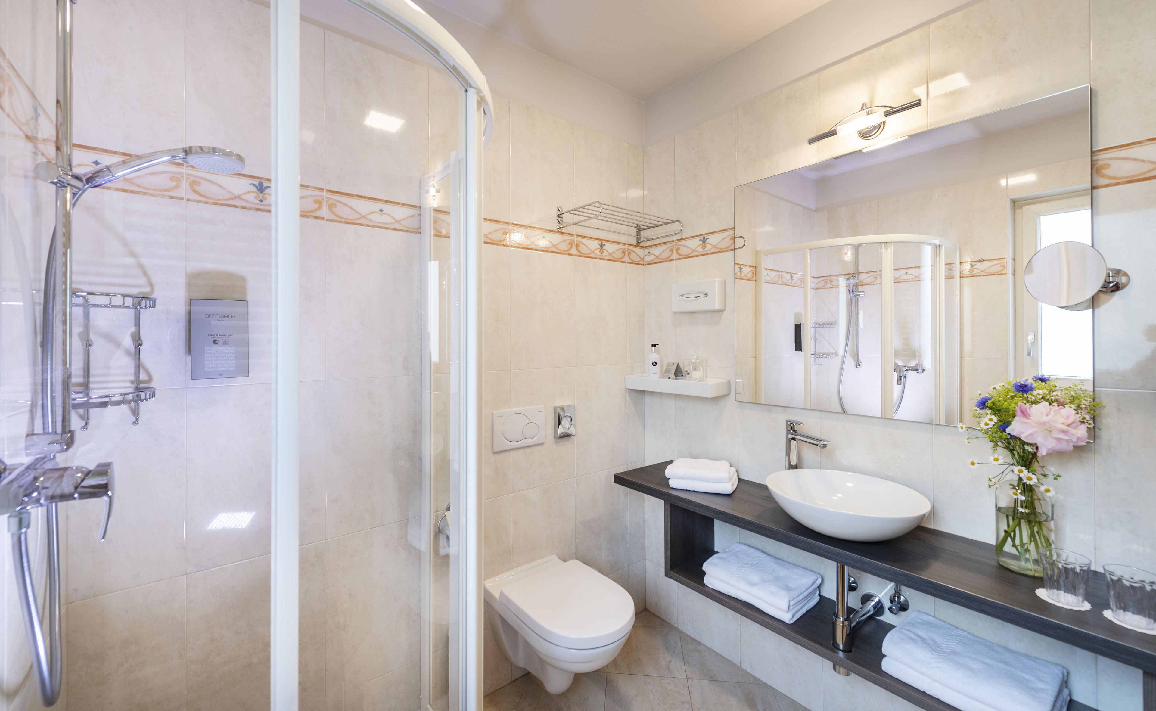 A modern bathroom with a glass-enclosed shower, a white toilet, a sink with a large mirror, and a vase with flowers on the countertop.