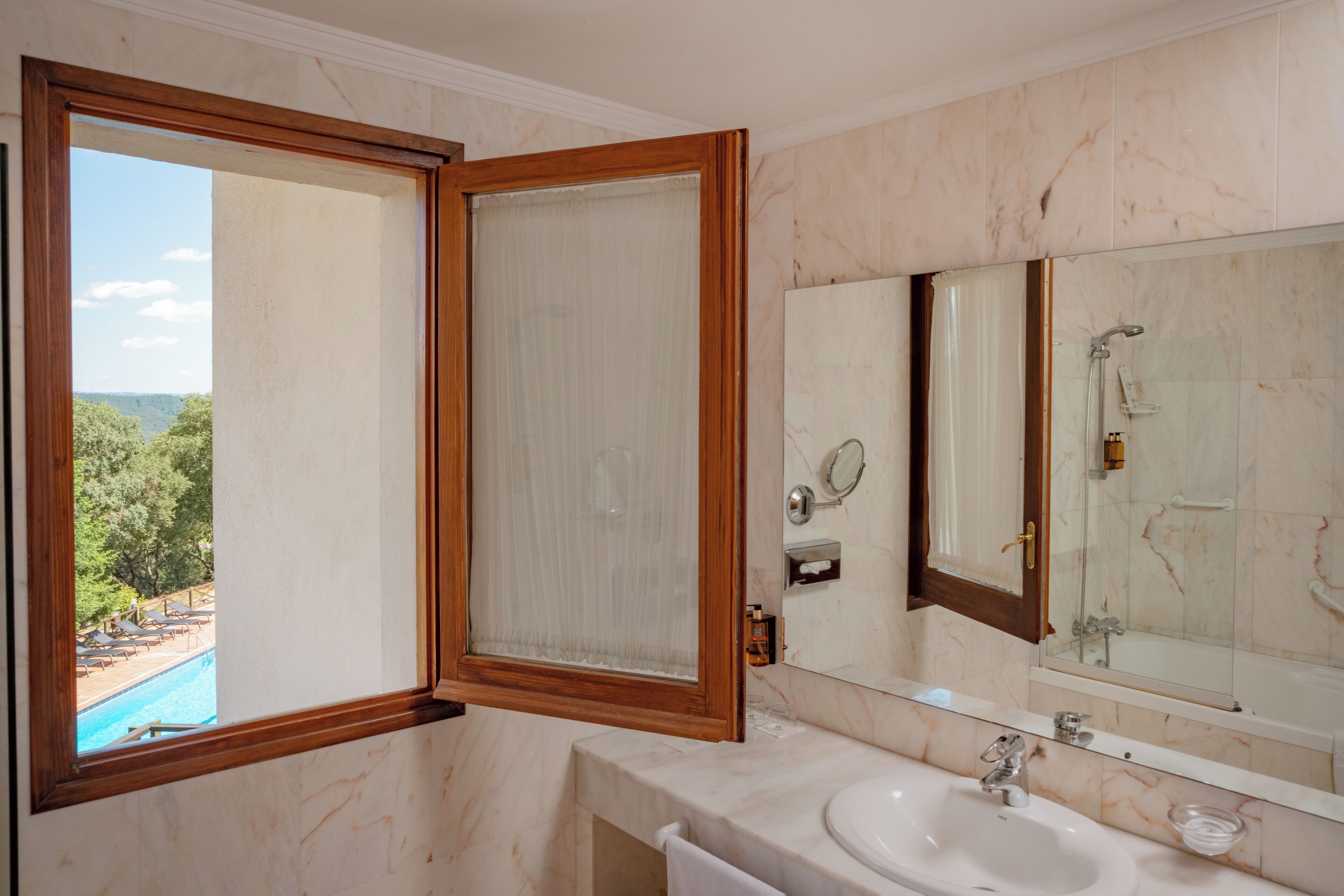 A bathroom with a large mirror, a sink, and a window with a view of a pool and trees.