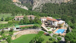 Un resort con varias construcciones, una piscina y una cancha de tenis, todo rodeado de follaje y montañas.