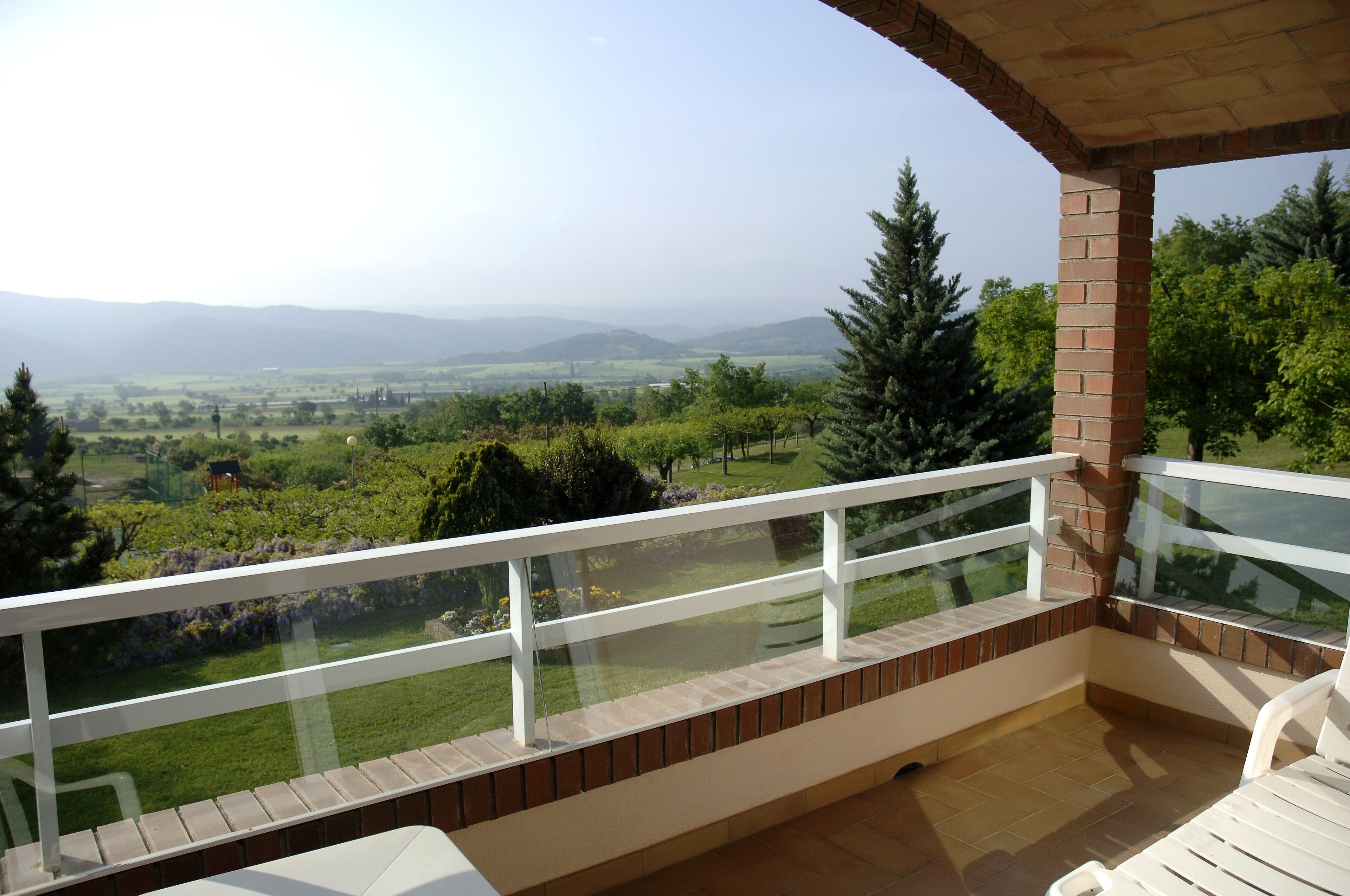 A balcony with a glass railing overlooking a green landscape and distant mountains.