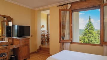 Ein Hotelzimmer mit Bett, Fernseher auf einer Holzkommode, einem Fenster mit Blick auf Bäume und einem Sessel in der Ecke.