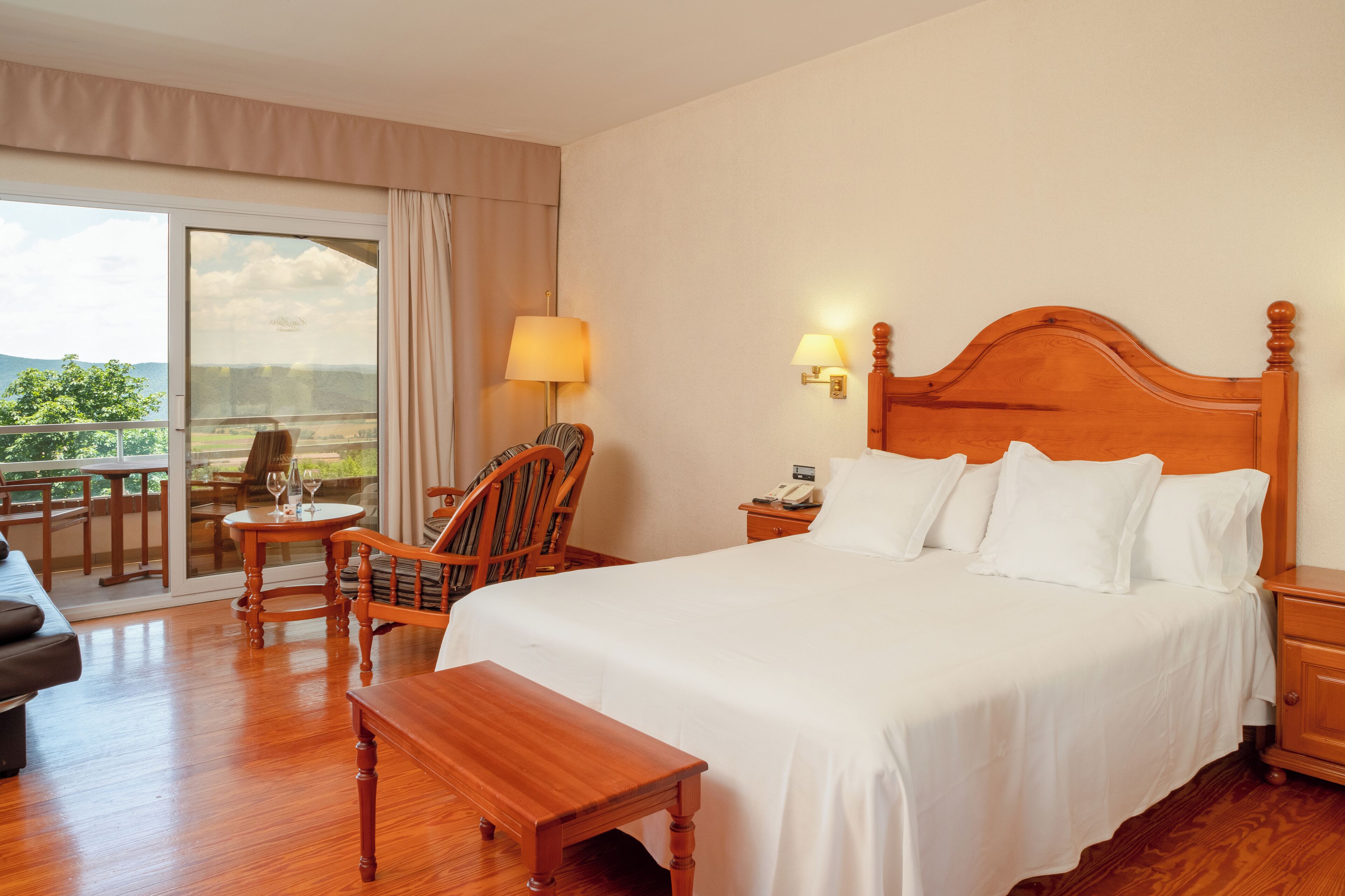 A hotel room with a large bed, a wooden nightstand, a chair, a small table with a lamp, and a view of the outdoors through a sliding glass door.