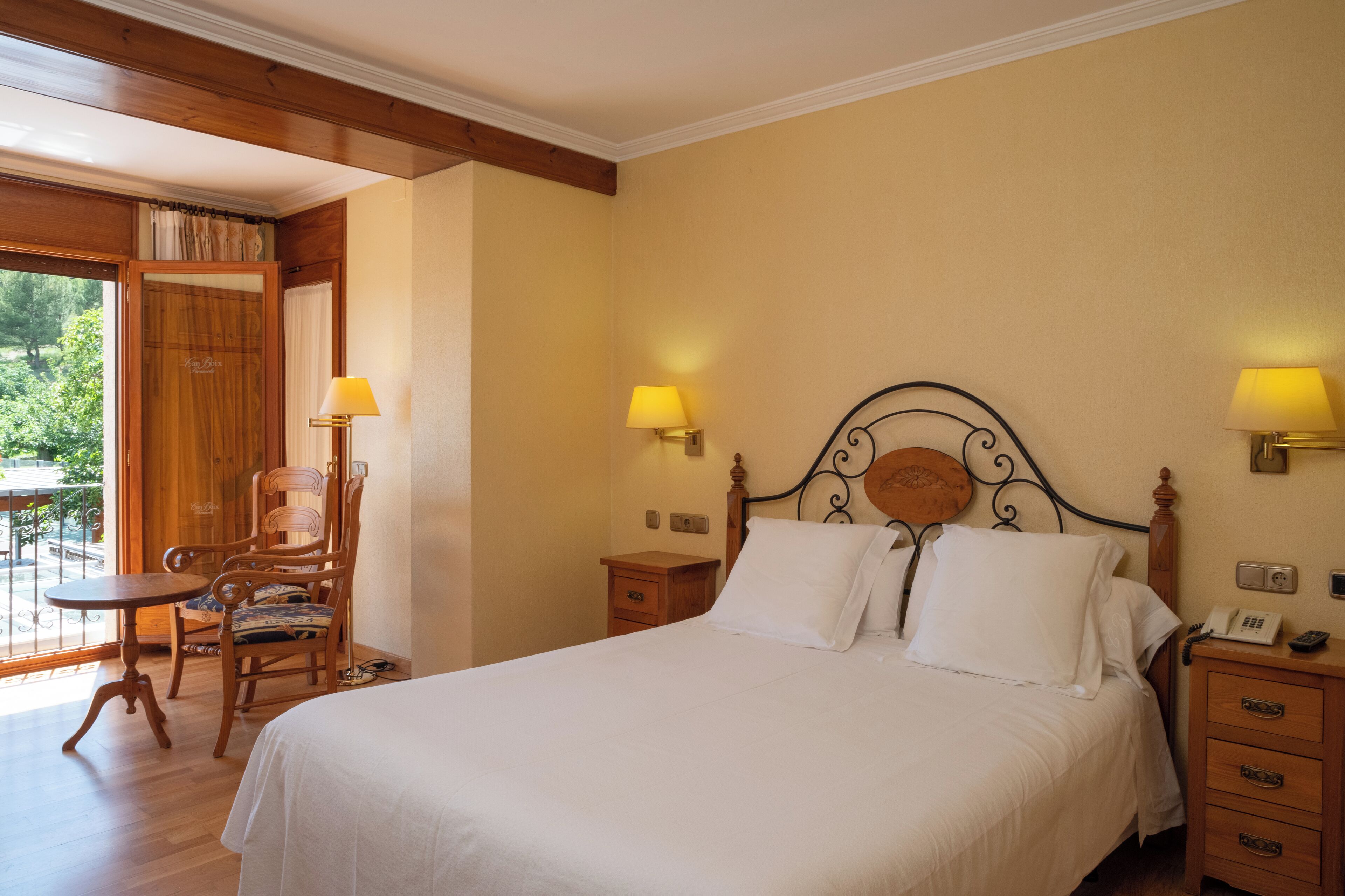 A hotel room with a bed, two bedside tables with lamps, a wooden chair, a small table, and a view of the outdoors.
