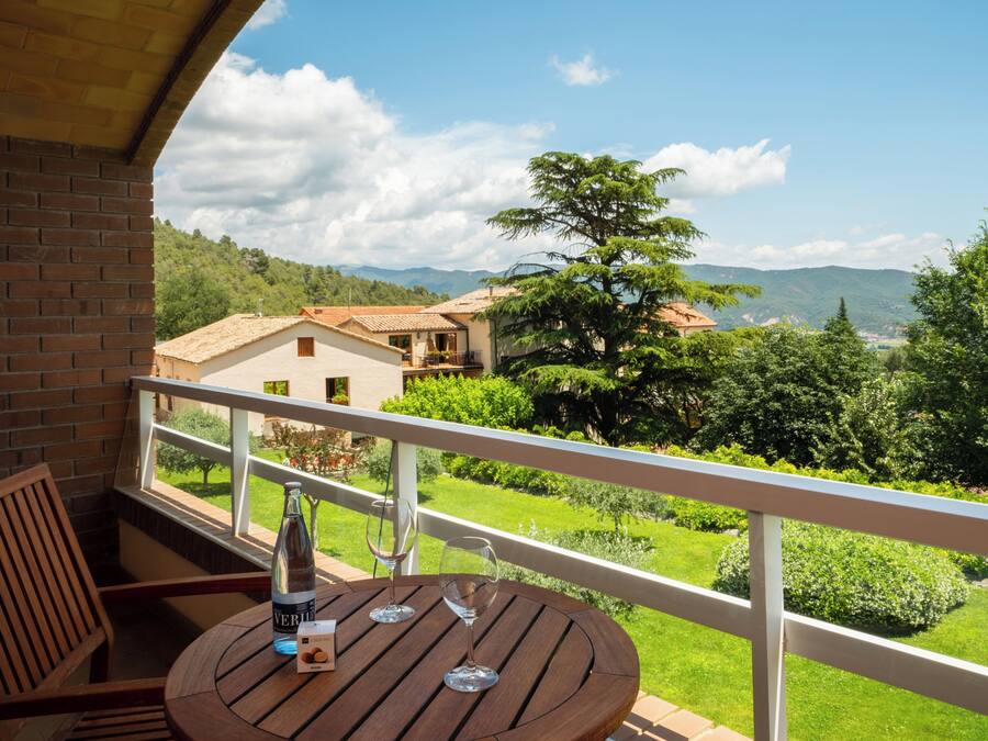 A balcony with a wooden table, two wine glasses, and a bottle. The balcony overlooks a lush garden and distant hills.