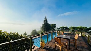 Terraza de madera con piscina, rodeada de follaje y cielo despejado.