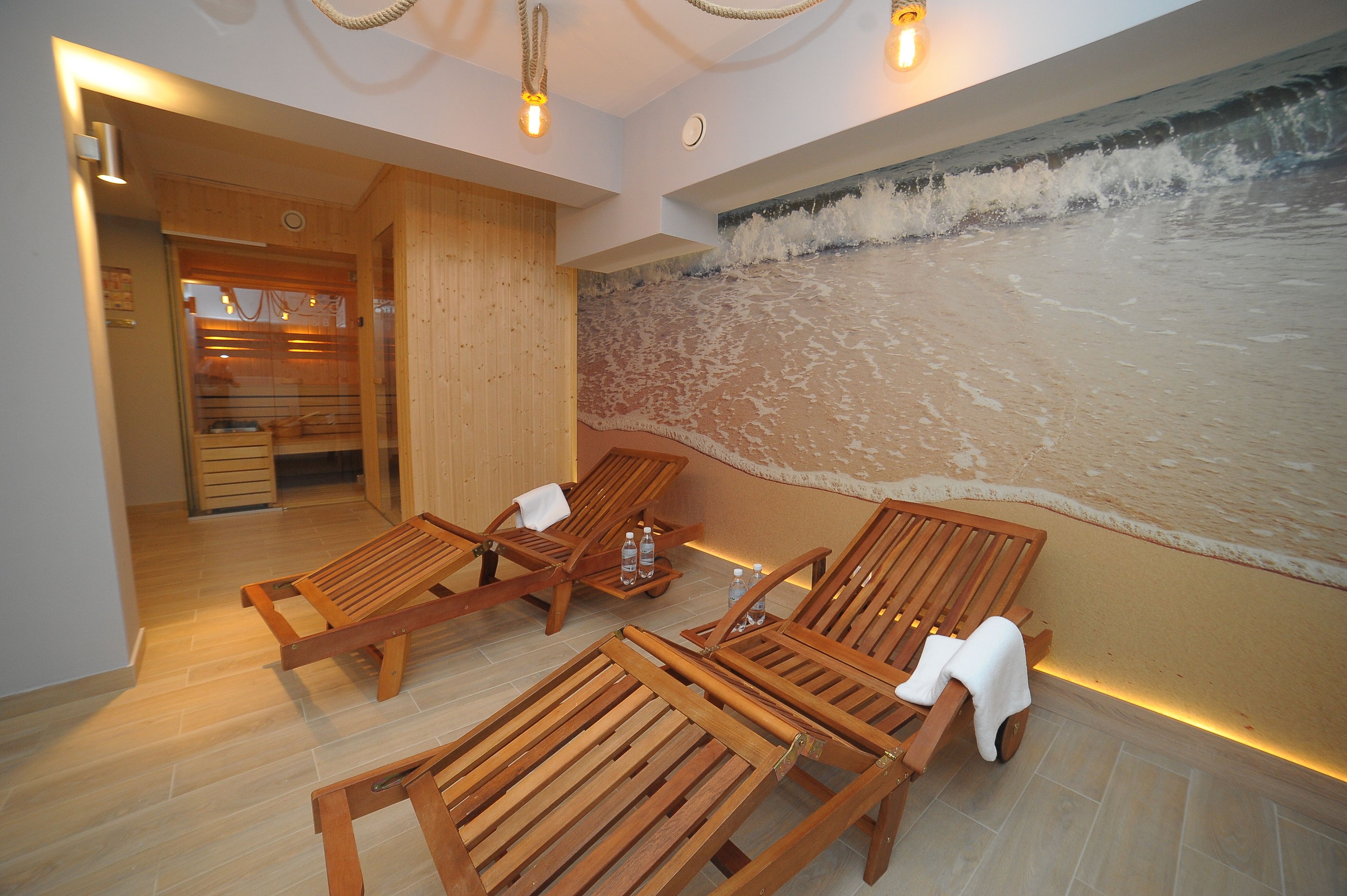 A spa area with wooden loungers, a wall mural of waves, and a wooden door leading to a room with a glass shower.