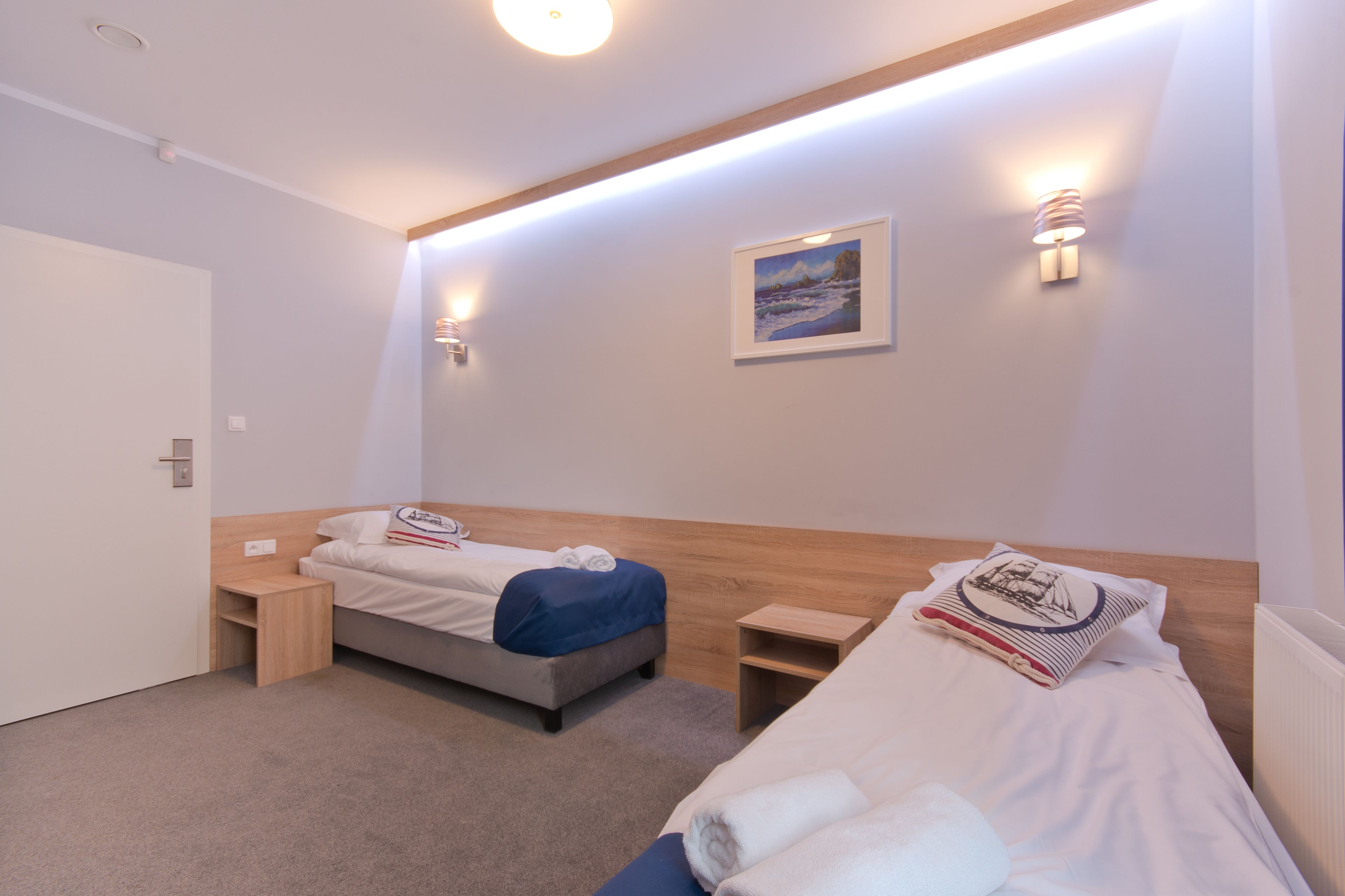 Une chambre d’hôtel avec deux lits simples, une petite table de chevet en bois, un tableau accroché au mur et un éclairage intégré au plafond.