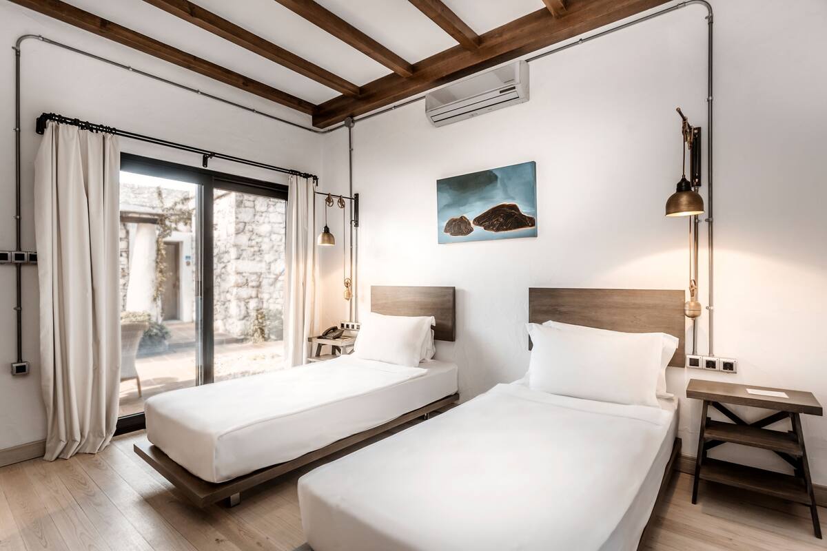 A hotel room with two beds, a large window with curtains, a painting on the wall, and a bedside lamp.