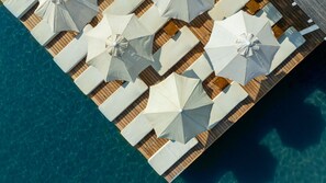 An aerial view of a wooden deck with white lounge chairs and umbrellas, overlooking clear blue water.