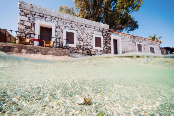 A stone building with multiple doors and windows, a balcony with wicker chairs, and a clear pool with a shell on the floor.