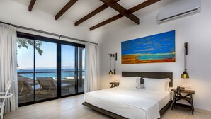 Un dormitorio con una cama grande, un cuadro de una playa, vista al mar a través de una puerta corredera y una mesita de noche con lámpara.