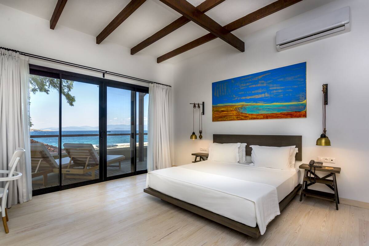 A bedroom with a large bed, a painting of a beach, a view of the sea through a sliding door, and a small table with a lamp.