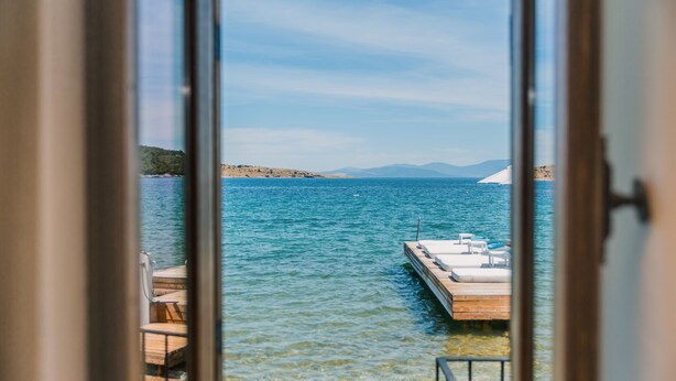 Vista a través de una ventana de un muelle de madera que se adentra en un mar tranquilo, con cielo despejado y tierra a lo lejos.