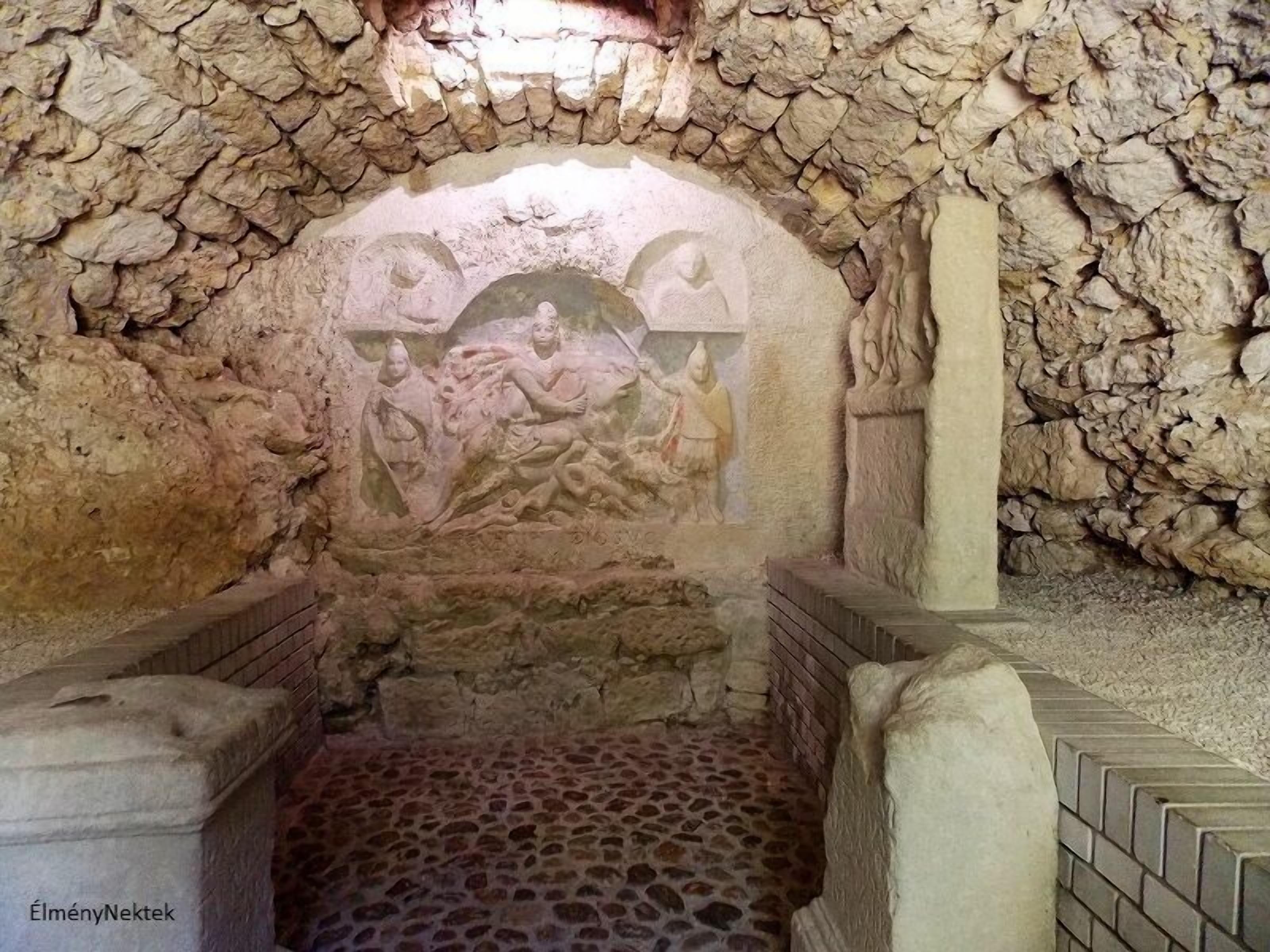 A stone-walled room with a central carved relief depicting figures, a stone floor, and a brick structure at the entrance.