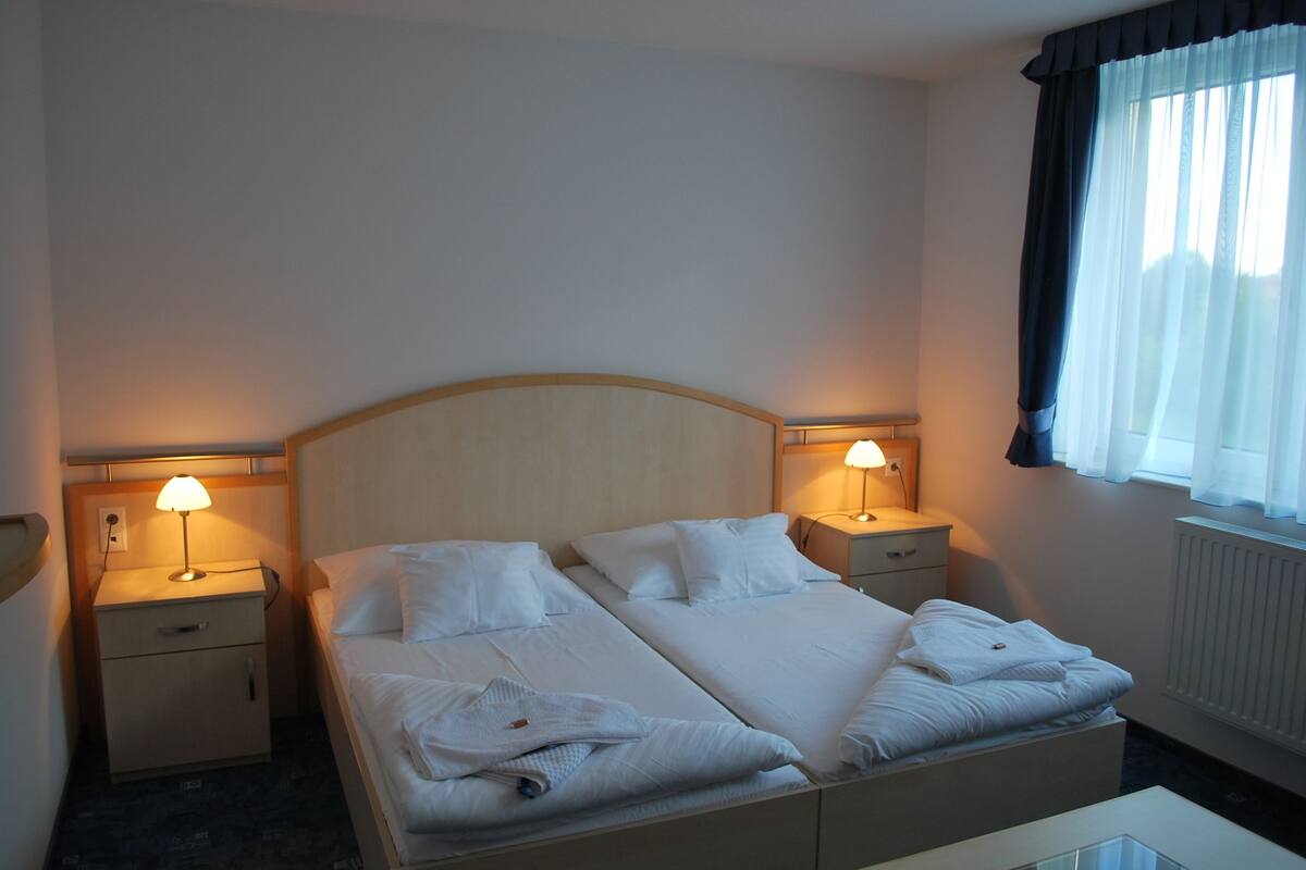A double bed with white linens, two bedside tables with lamps, a window with curtains, and a radiator.