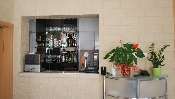 A bar area with a coffee machine, a cash register, and a variety of bottles and glasses. There is a potted plant with red flowers on a small table. - Főnix Hotel Bükfürdő (Buk)
