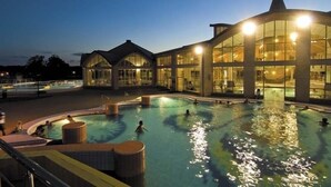A swimming pool area with a large building in the background, including a covered section and a tower-like structure. - Főnix Hotel Bükfürdő (Buk)