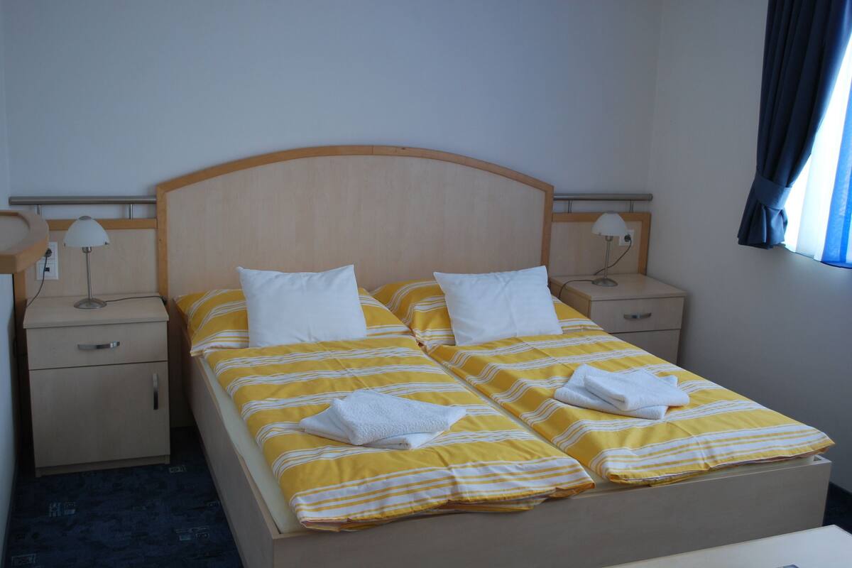A double bed with yellow and white striped bedding, two white pillows, and a white towel folded on the bed. There are two bedside tables with lamps, a wooden headboard, and a window with blue curtains.