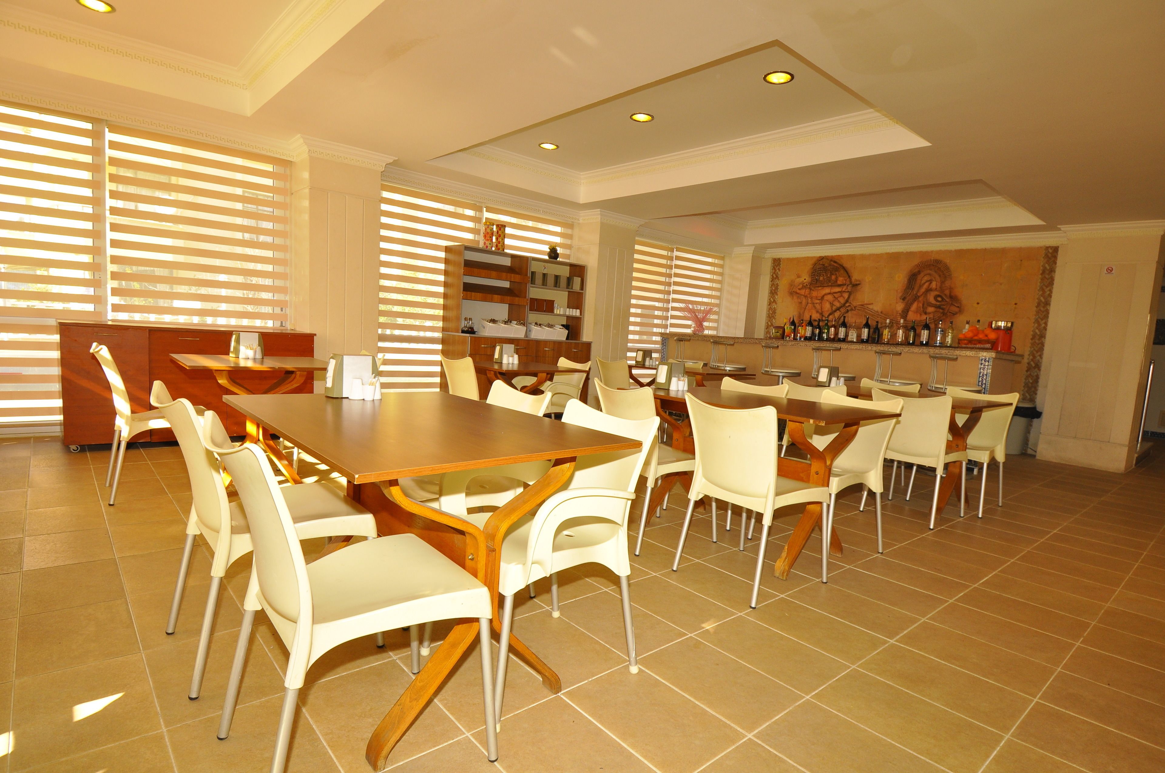 A spacious dining area with a long wooden table and white chairs, a bar with various bottles, and a large window with blinds.