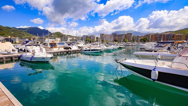 Un puerto deportivo con barcos amarrados, rodeado de edificios y montañas al fondo.