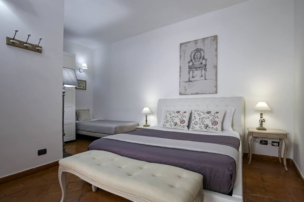 Quarto com uma cama grande, duas mesas de cabeceira com luminárias, um banquinho, uma cadeira e uma obra de arte na parede.