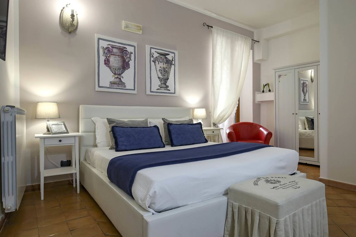 Quarto bem arrumado com cama, mesas de cabeceira, uma cadeira vermelha e um guarda-roupa.