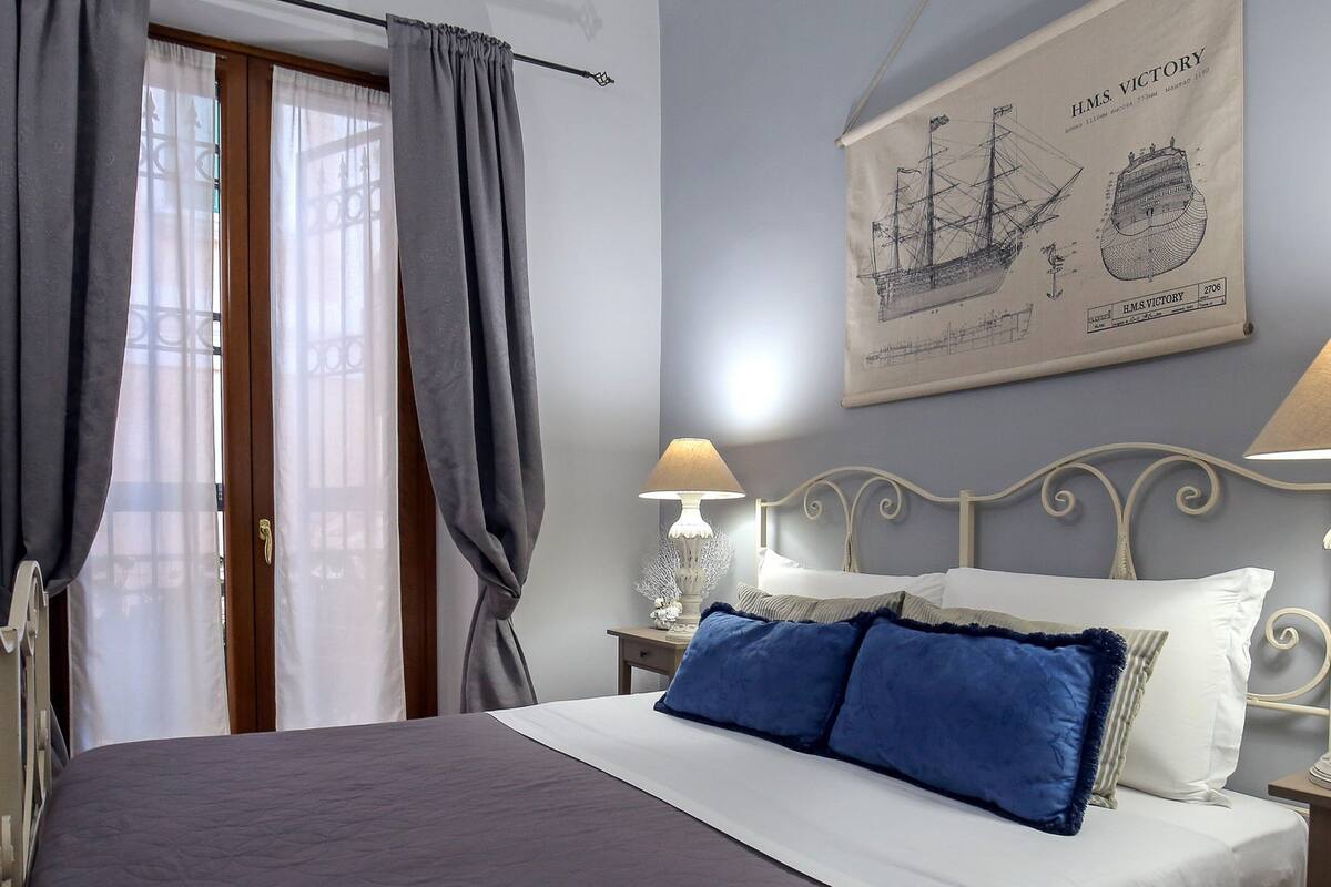 Quarto com cama, cortinas, luminária na parede e quadro de um navio.