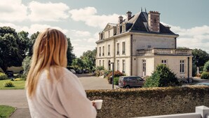 A woman standing on a balcony overlooking a large, historic building with a symmetrical facade and a central entrance. - Château de la Gressière (La Bernerie-en-Retz)