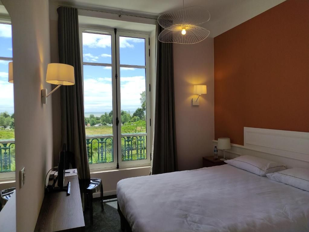 A hotel room with a bed, a desk, a chair, a window with curtains, and a view of greenery.