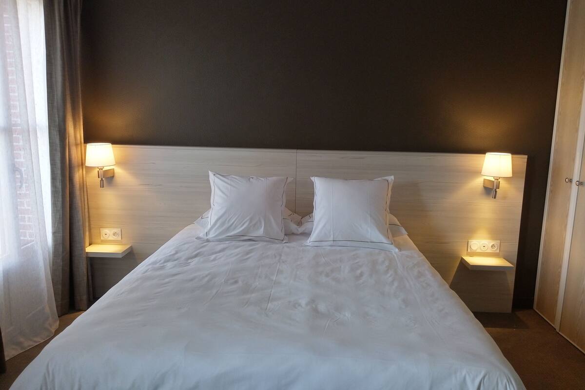 A hotel room with a large bed, two bedside lamps, and a plain headboard.