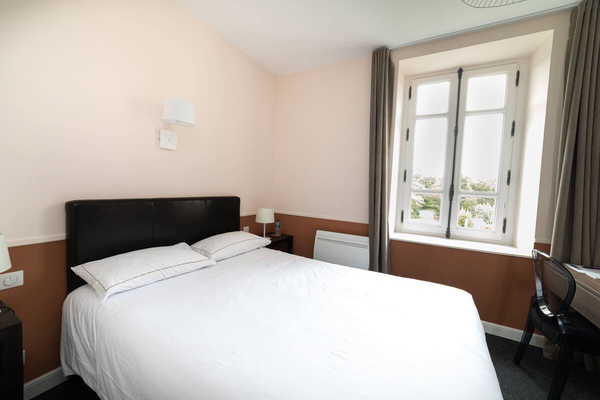 A hotel room with a large bed, a nightstand with a lamp, a chair, and a window with curtains.