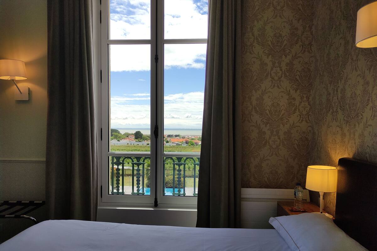 A hotel room with a large window, a bed, a nightstand, and a lamp.