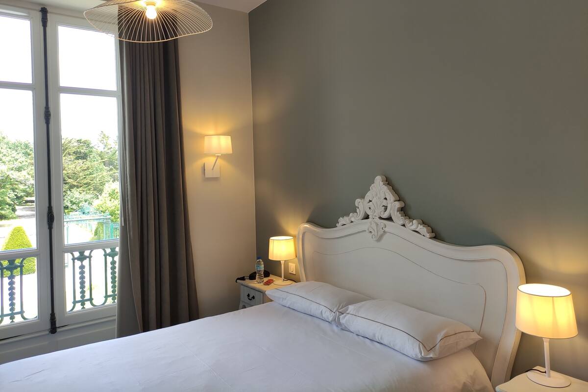 A hotel room with a bed, two bedside lamps, a window with curtains, and a view of greenery outside.