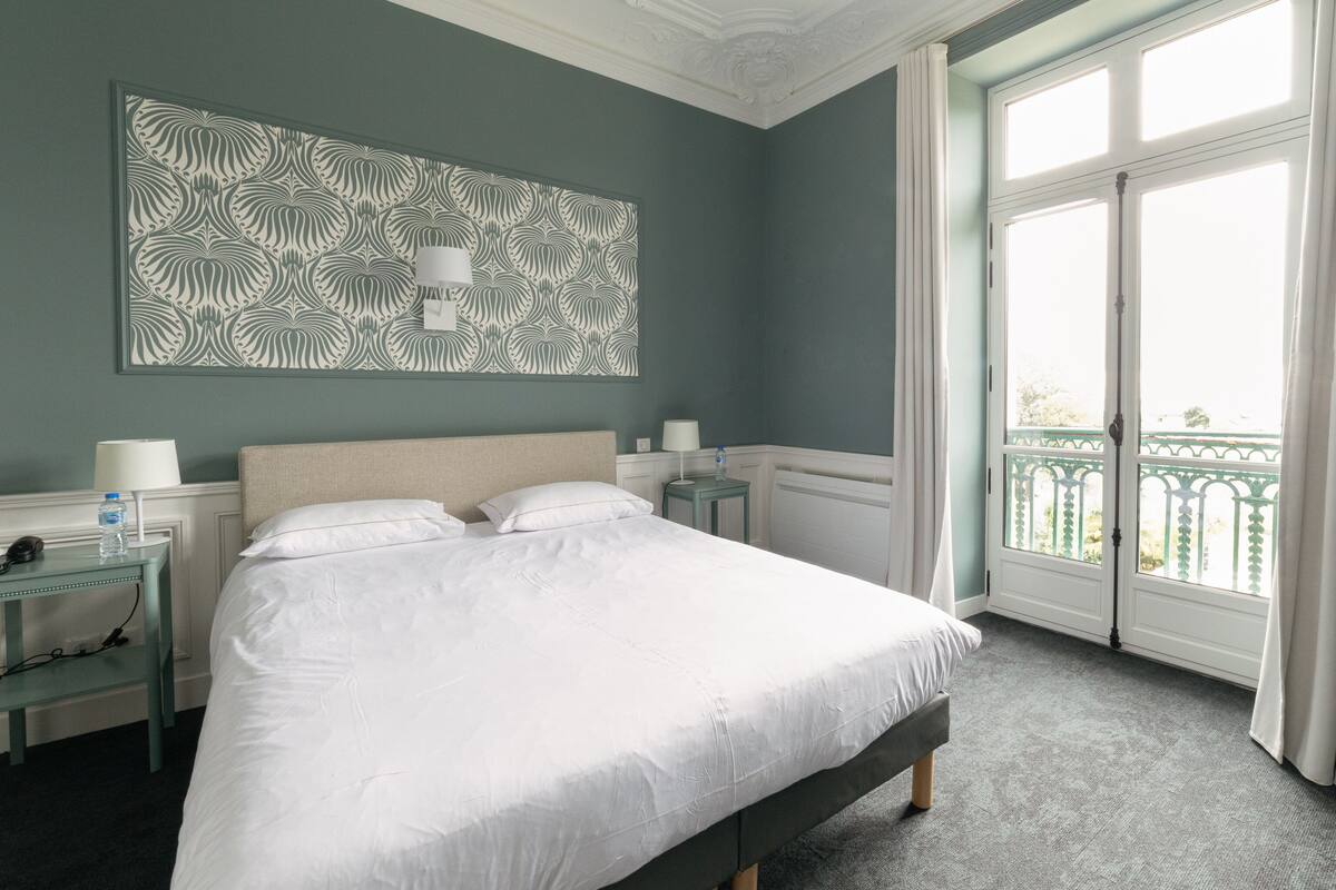 A neatly made bed in a hotel room with a large window, a decorative wall piece, and a small bedside table with a lamp.