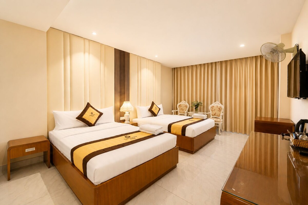 A hotel room with two beds, a desk, a chair, a TV, and a large window with curtains.
