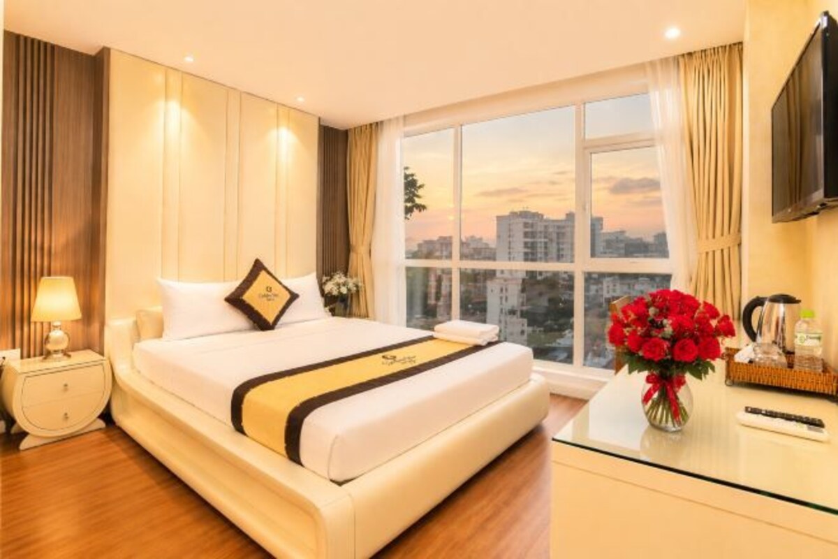 A hotel room with a bed, a nightstand, a vase of flowers, a TV, and a window with a city view.