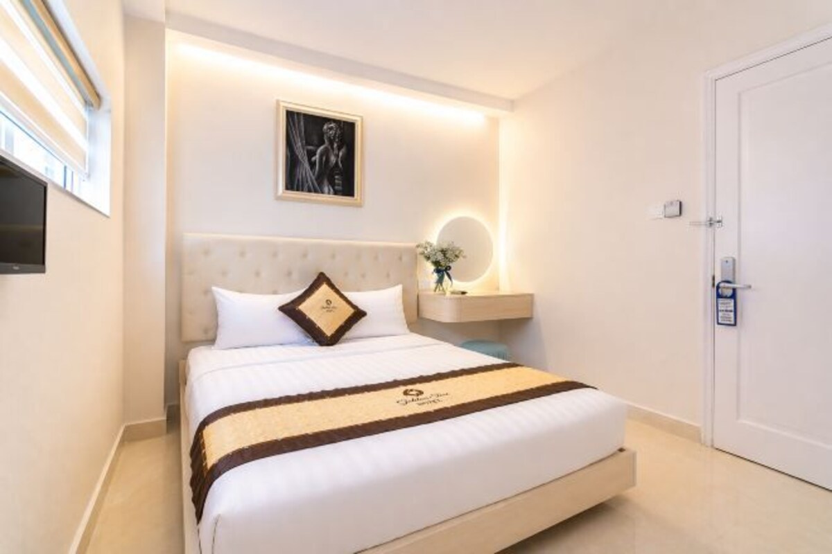 A hotel room with a bed, a television, a wall-mounted artwork, a bedside table with a vase, and a door.