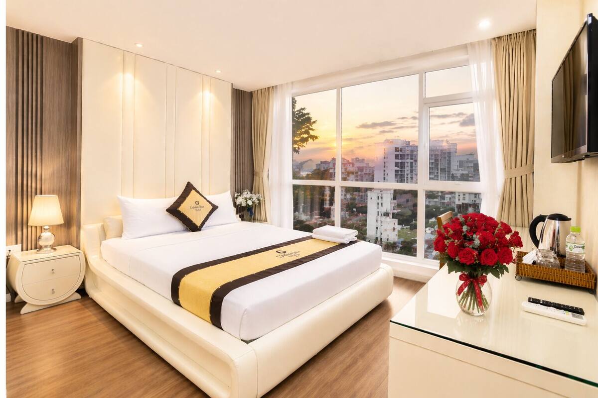 Een hotelkamer met een groot bed, een nachtkastje, een televisie, een raam met uitzicht op de stad en een vaas met bloemen.